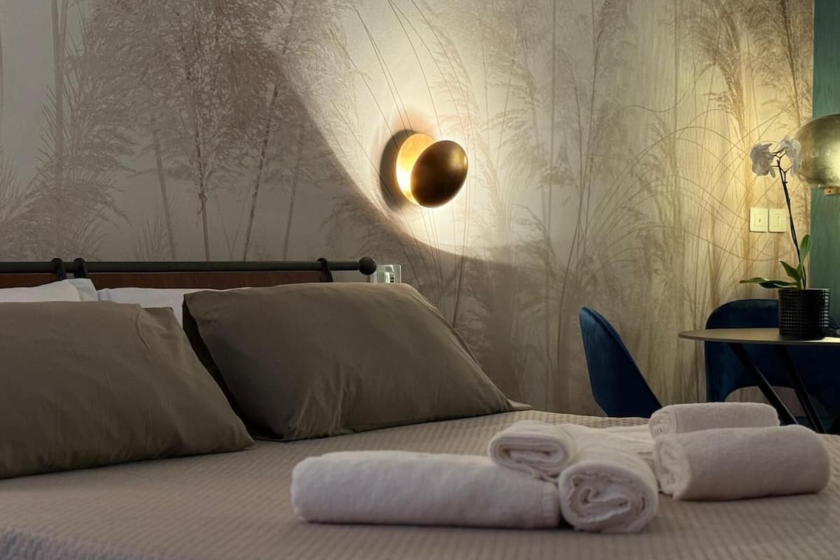 Ein ordentlich bezogenes Bett mit Kissen, einer Wandleuchte und einem kleinen Tisch mit einer Pflanze.