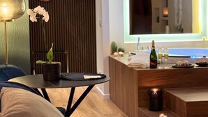 A modern hotel room with a wooden floor, a round table with a plant, a sofa, a large mirror with an arch, and a bar area with a bottle and glasses. - Via del Mare Apulia B&B  Spa Suite (Rutigliano)