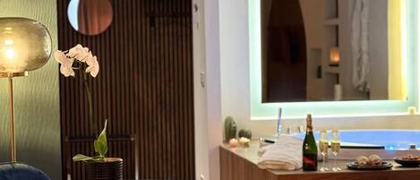 Una habitación de hotel moderna con piso de madera, una mesa redonda con una planta, un sofá, un espejo grande de arco y una zona de bar con una botella y vasos.
