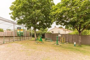 A fenced outdoor area with playground equipment, trees, and a building in the background.