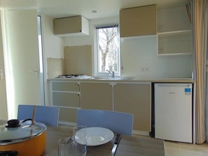 Cocina funcional con estufa a gas, lavamanos y refrigerador. Cuenta con una mesa de comedor con sillas y un ventanal que proporciona luz natural.