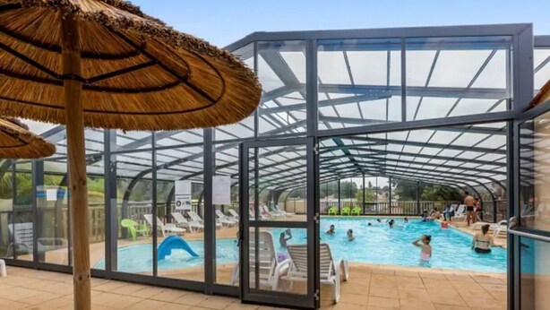 Une piscine intérieure avec des parois en verre transparent, des chaises longues et un parasol en chaume.