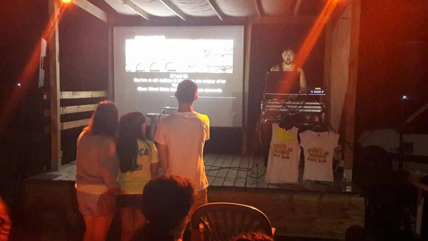 An outdoor event with a stage, a projector screen, and a DJ setup.