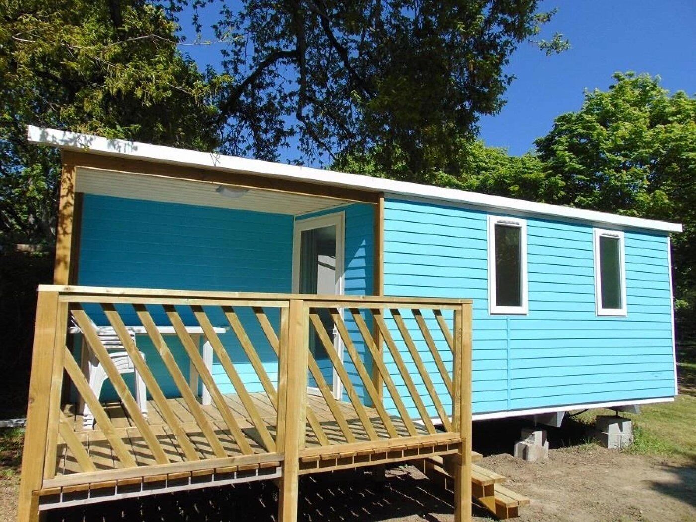 A small, single-story mobile home with a blue exterior, a wooden deck, and a window on the side.
