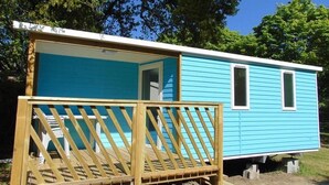 Une petite maison mobile d’un seul étage, à l’extérieur bleu, avec une terrasse en bois et une fenêtre sur le côté.
