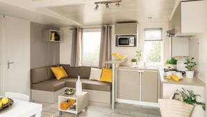 A compact living space with a sofa, a small dining table, a kitchenette, and a window with curtains. - ZEcamping 3* piscine couverte locatif TV LV 31m2 (Avrillé)