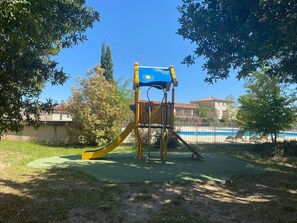Un terrain de jeux avec un toboggan et une structure d’escalade, entouré d’arbres et avec une piscine en arrière-plan.