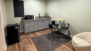 Una cocina compacta equipada con refrigerador, estufa y microondas. Hay una mesa de comedor con sillas y una alfombra pequeña. El piso es de madera y las paredes son de color verde claro.