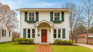 A two-story house with a red front door, green shutters, and a brick pathway leading to the entrance.