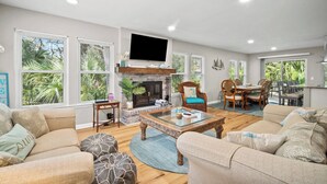 A spacious living room with a fireplace, comfortable seating, a dining area, and large windows with views of greenery. - Frippin Graytful, Golf Cart Included, Pet Friendly - 120 Sea Otter (Fripp Island)