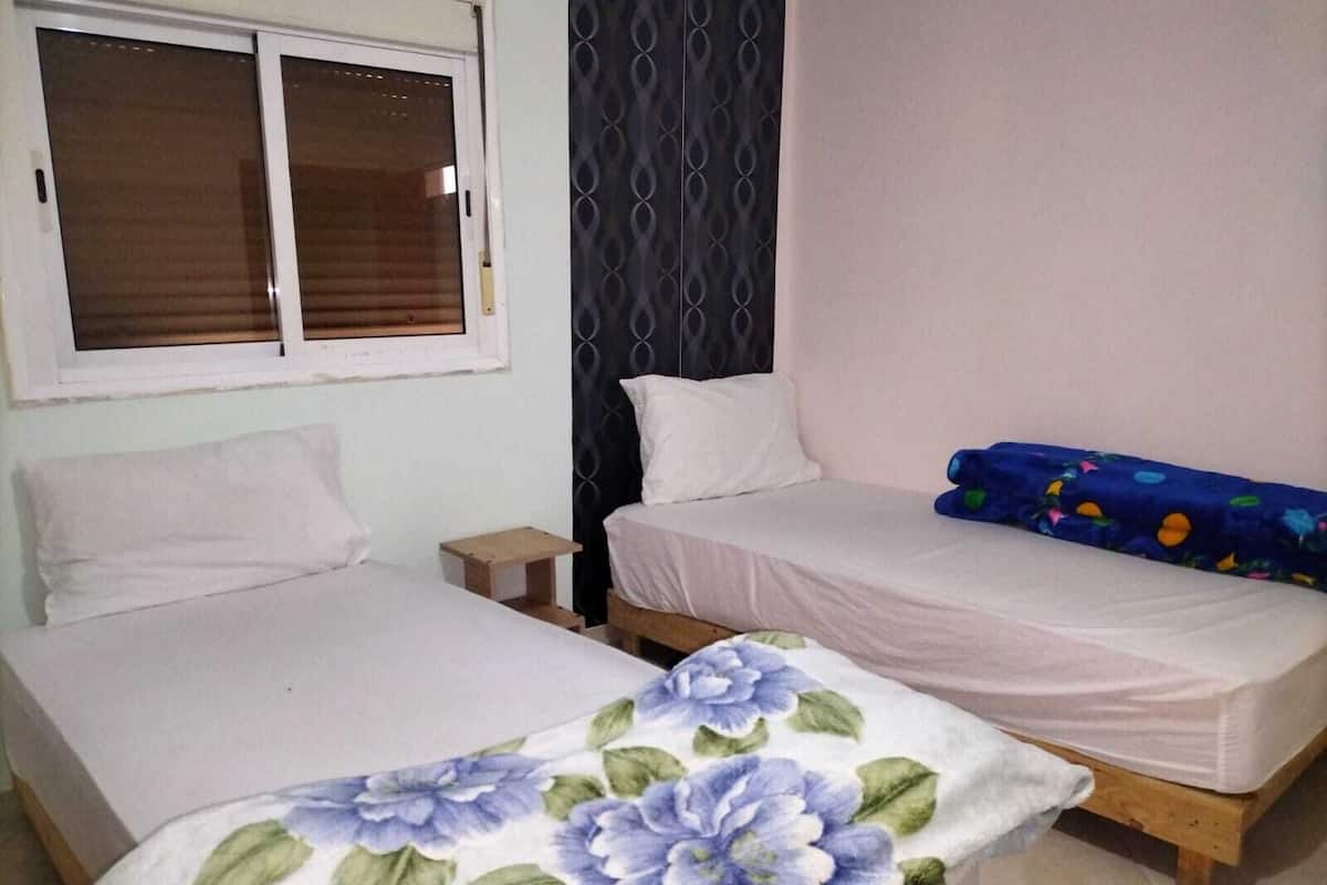 A room with two beds, a window with blinds, a small wooden nightstand, and a patterned bedspread.