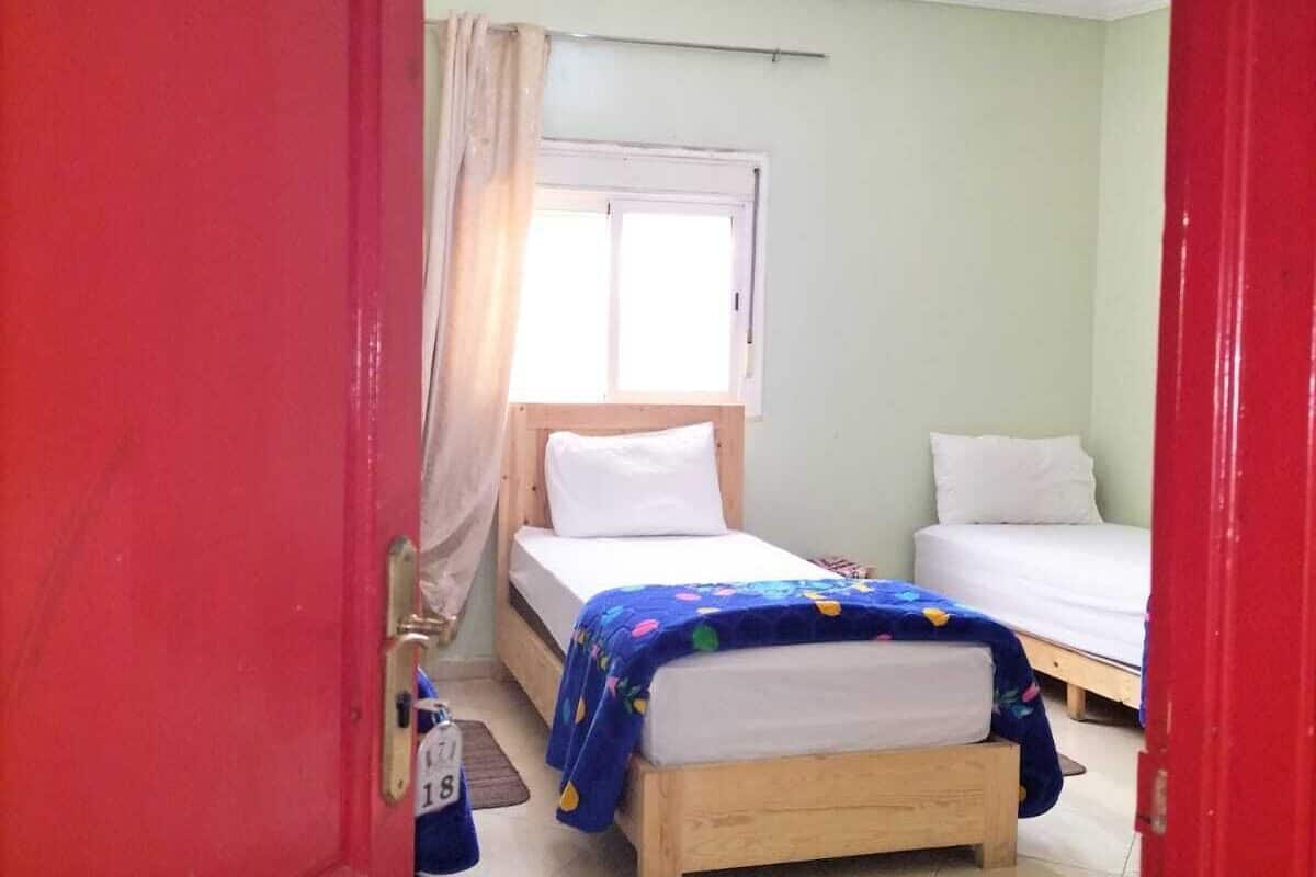 A room with two beds, a window with curtains, a ceiling light, and a red door.