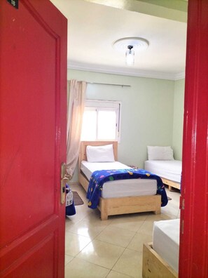 A room with two beds, a window with curtains, a ceiling light, and a red door. - HOTEL 7DAYS (Dakhla)