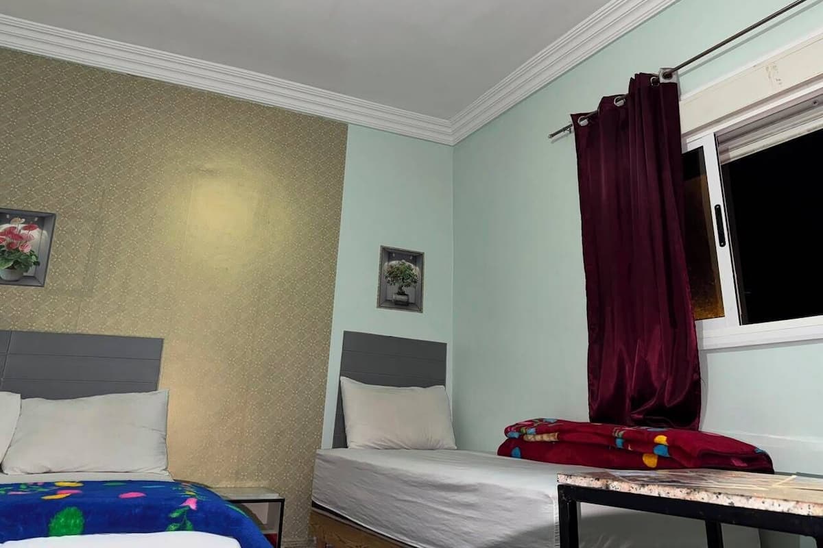 A bedroom with two beds, a window with curtains, a small table, and a wall-mounted picture.