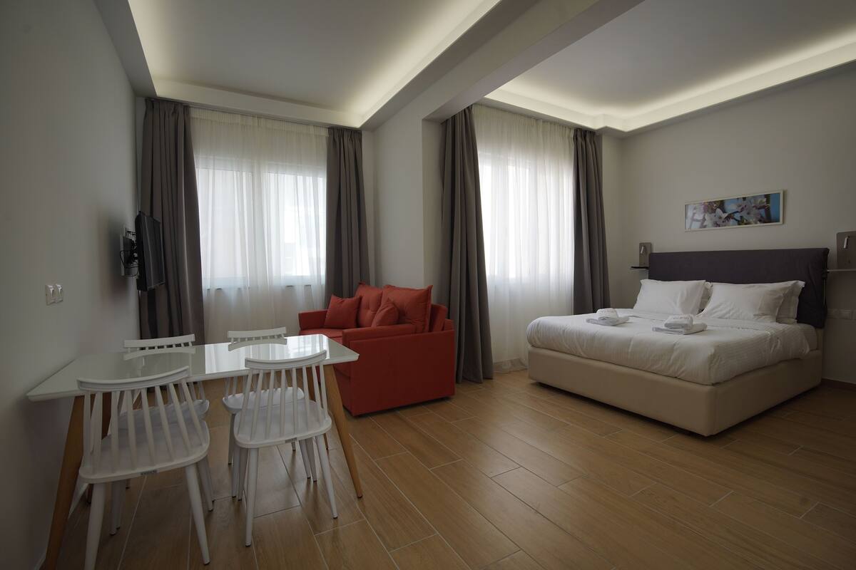 A modern hotel room with a bed, a red sofa, a dining table with chairs, and a TV.