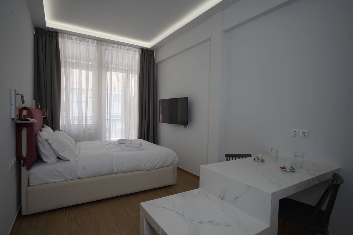 A modern hotel room with a bed, a TV mounted on the wall, a small table with a chair, and a window with curtains.