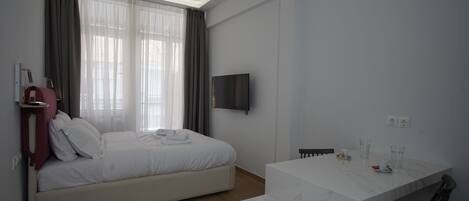 A modern hotel room with a bed, a TV mounted on the wall, a small table with a chair, and a window with curtains.