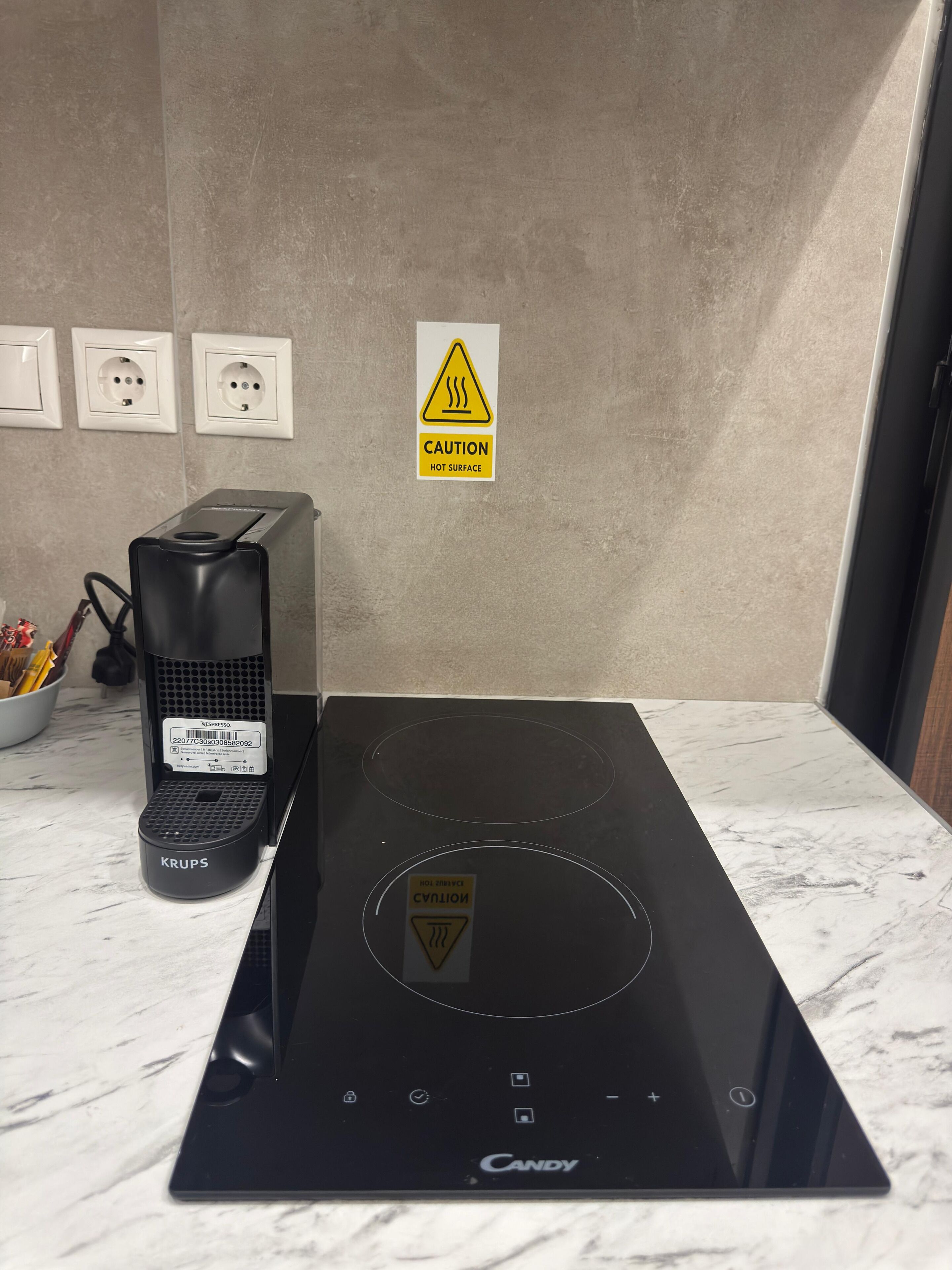 A kitchen with a black induction cooktop, a Krups coffee maker, and a caution sign for a hot surface.