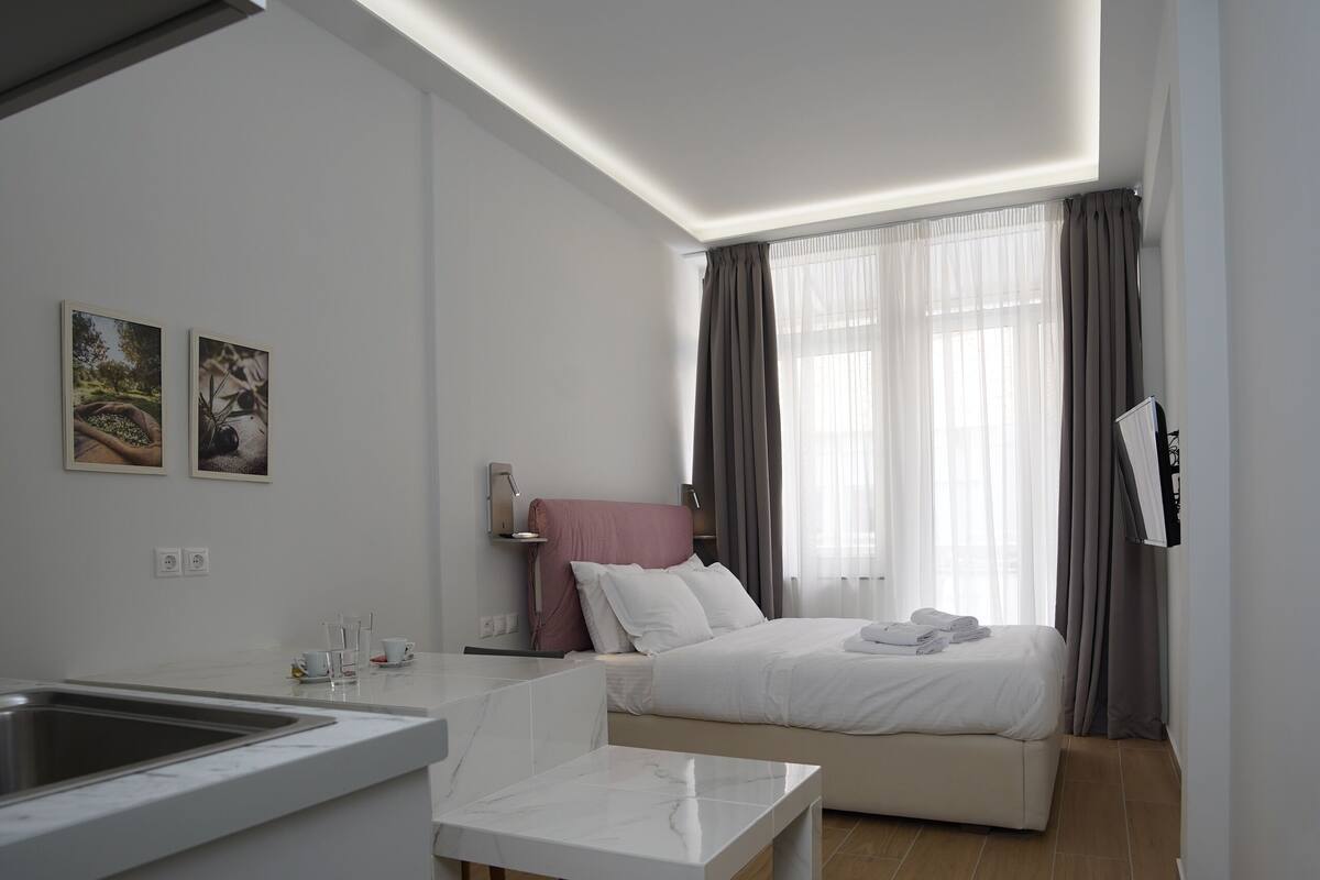 A modern, compact hotel room with a kitchenette, a bed with a pink headboard, and a large window with sheer curtains.