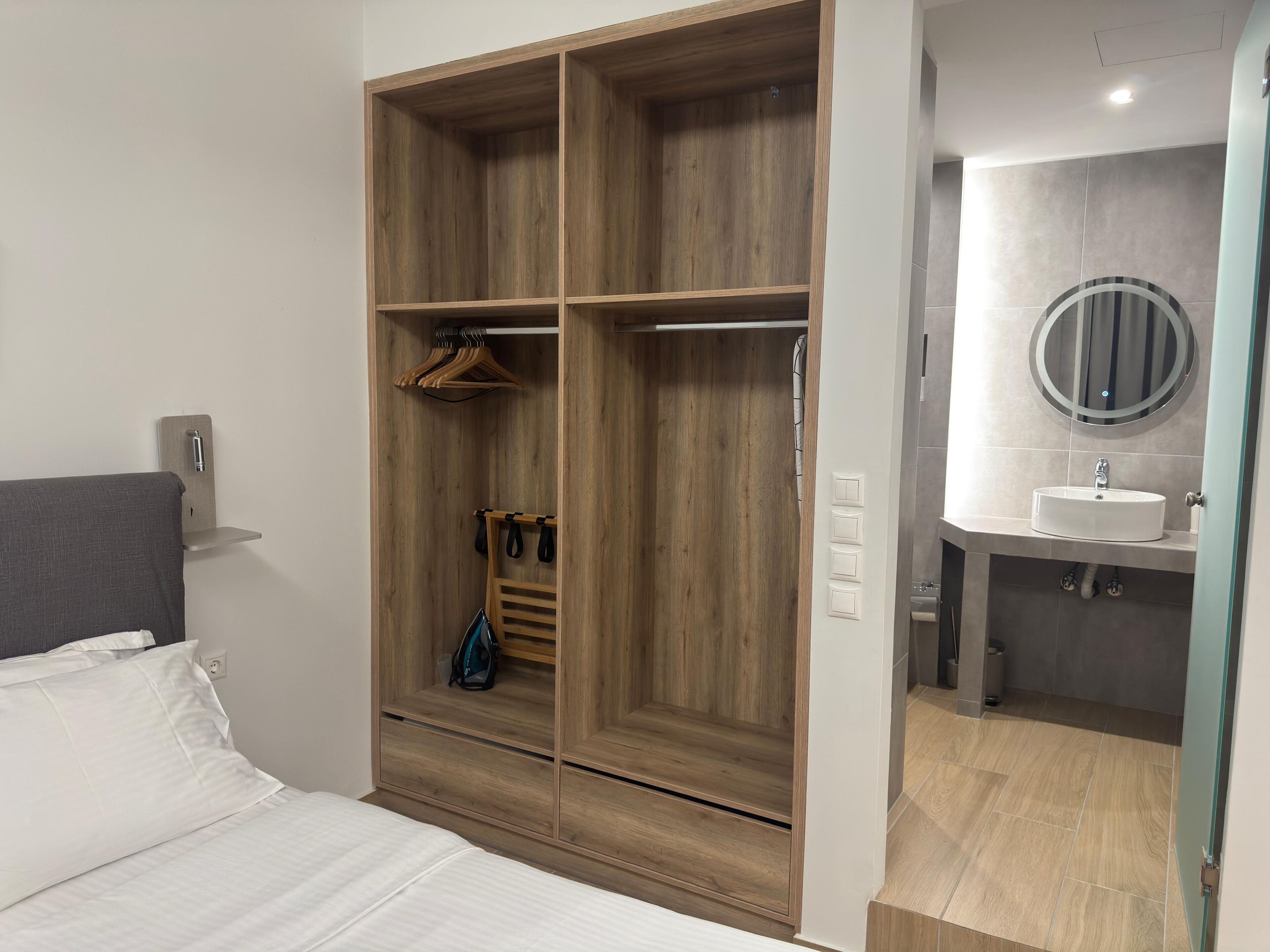 Un dormitorio con una cama, un armario de madera con varios compartimentos, un perchero y un baño con lavabo y espejo.