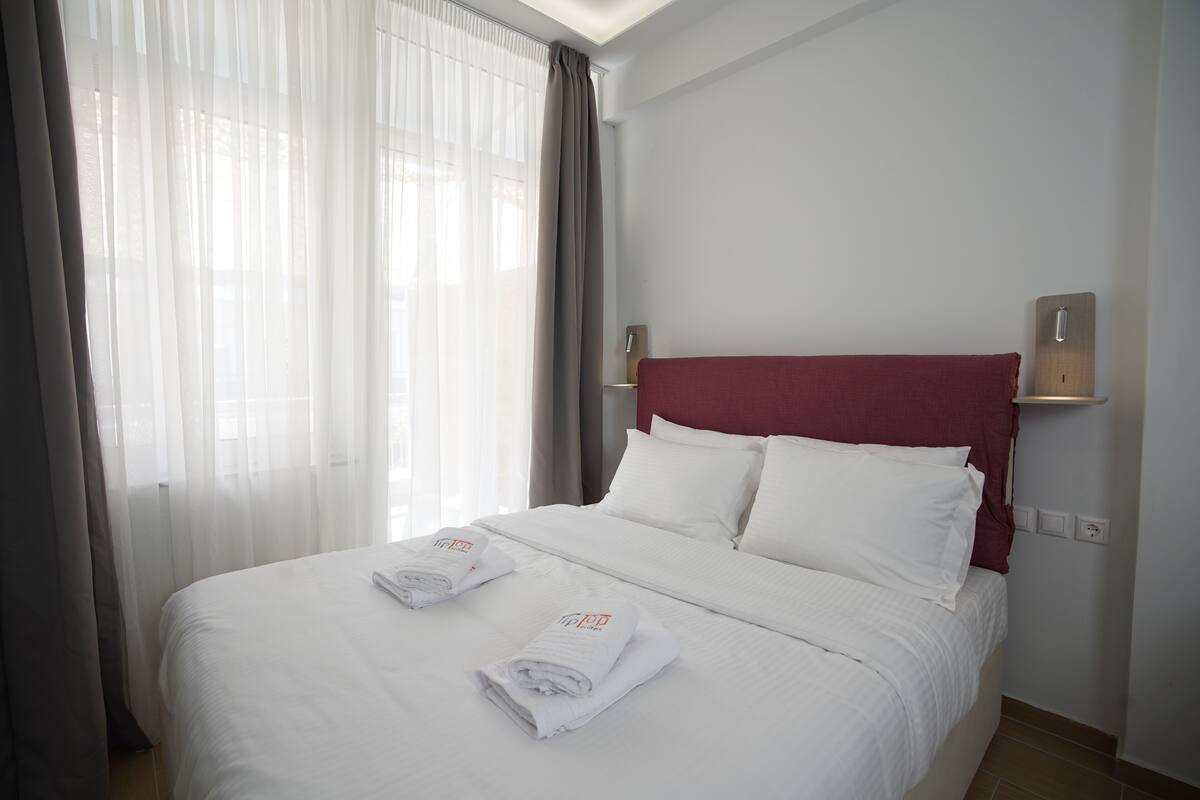 A hotel room with a neatly made bed, a red headboard, and a large window with sheer curtains.