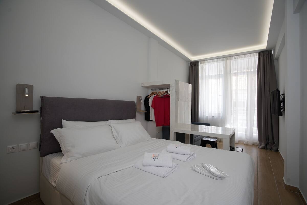 A modern hotel room with a neatly made bed, a bedside table, a desk, a wardrobe, and a large window with curtains.