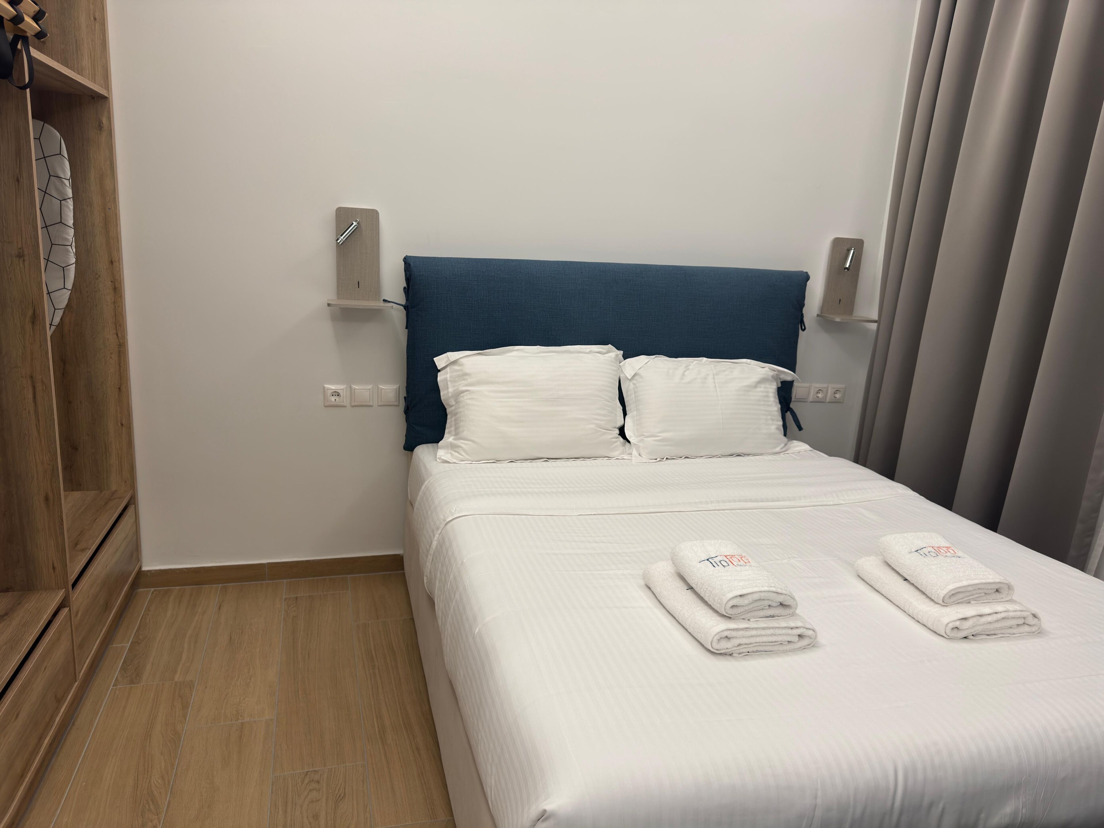 Una cama bien hecha con ropa blanca y dos toallas dobladas, un armario de madera y un cabecero azul.