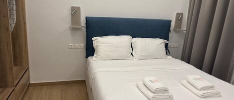 Una cama bien hecha con ropa blanca y dos toallas dobladas, un armario de madera y un cabecero azul.