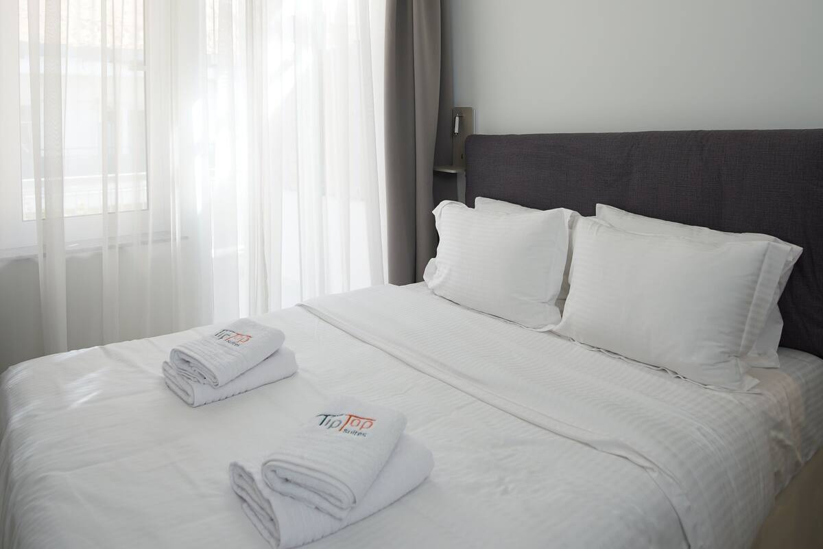 A neatly made bed with white linens and towels, a dark headboard, and a window with sheer curtains.