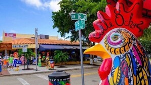 Una estatua colorida de un gallo en una esquina de calle con señales de tráfico y un restaurante al fondo.