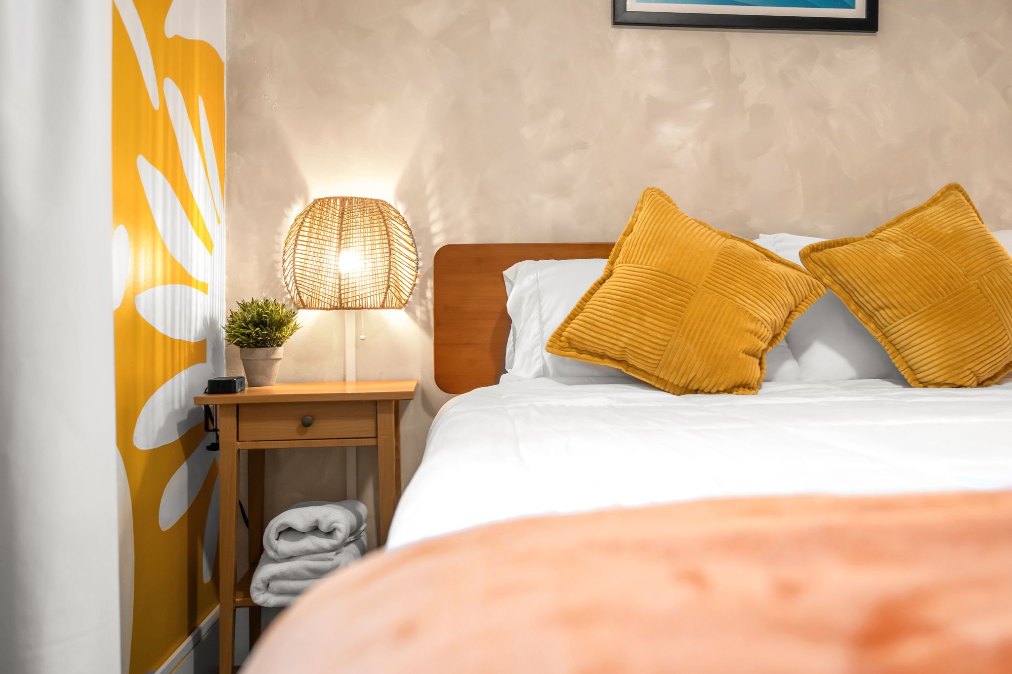 A neatly made bed with two pillows, a bedside lamp, a small plant, and a folded towel on a wooden nightstand.