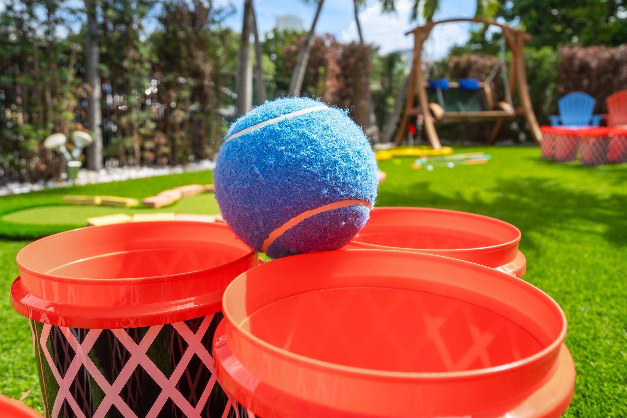 A blue ball on a red plastic table with a lattice base, set outdoors with greenery and a swing set in the background.