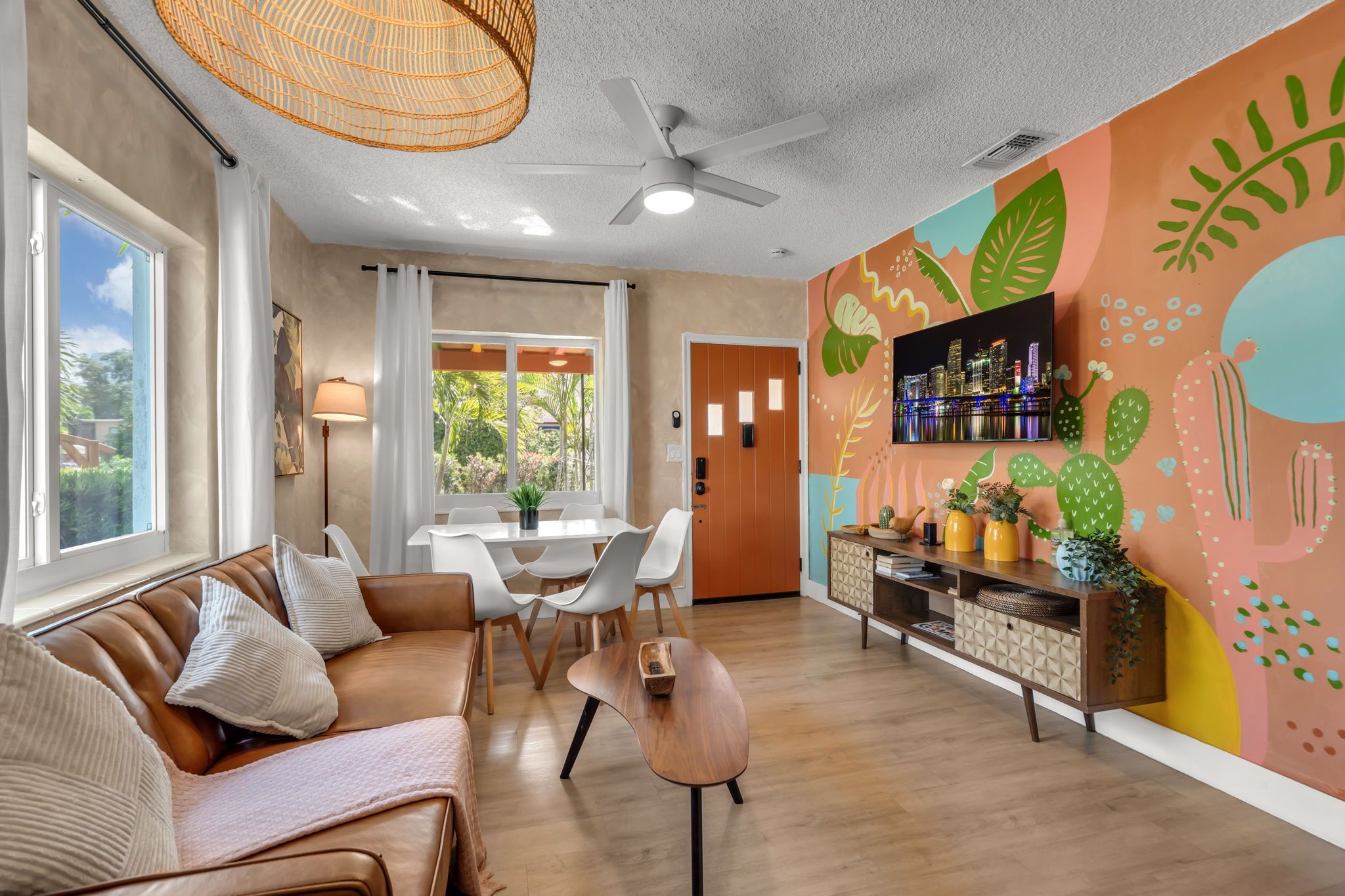 A modern living room with a sofa, dining table, and chairs, a ceiling fan, and a vibrant wall mural.