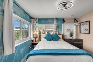 Quarto com cama, mesa de cabeceira, cômoda, ventoinha no teto e janela com cortinas.