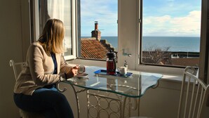 A person sitting at a glass table by a window with a view of the sea and a building with a chimney. - Ard na Mara- Coastal Apartment (Pittenweem)
