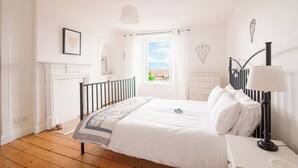 A bedroom with a bed, a nightstand, a lamp, a dresser, a window with curtains, a fireplace, and a heart-shaped decoration on the wall.