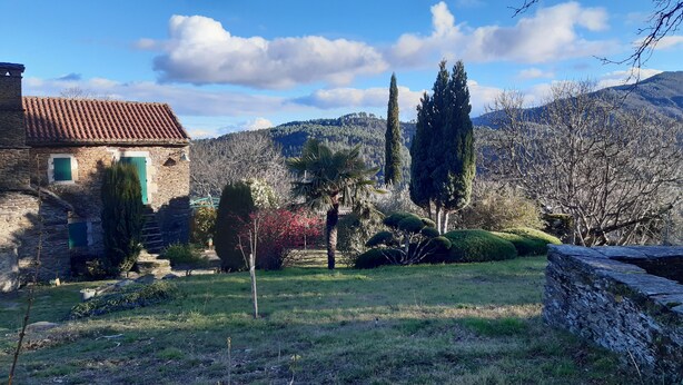 Un edificio de piedra con tejado de tejas, rodeado de un jardín bien cuidado con árboles y arbustos diversos, con montañas como telón de fondo.
