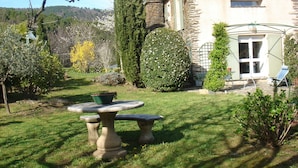 Un edificio de piedra con jardín que incluye una mesa de piedra, césped y diversas plantas.