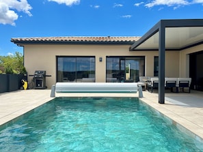 Uma área moderna ao ar livre com piscina azul cristalina, uma espreguiçadeira e uma churrasqueira.
