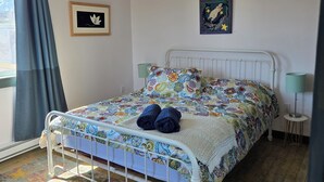A bedroom with a bed, a chandelier, a window with curtains, and a colorful rug. - Oceanfront Apartment, Gros Morne View in the heart of Park (Rocky Harbour)