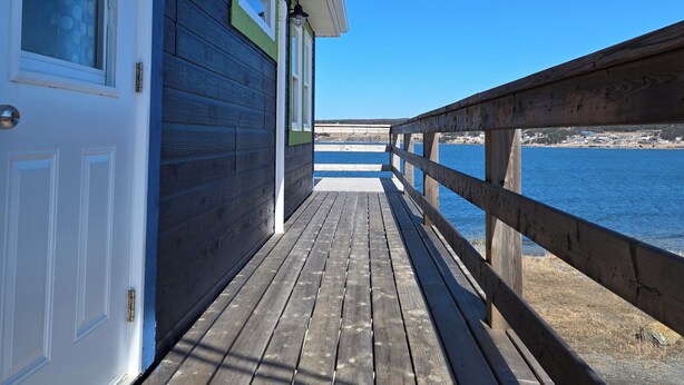 A wooden deck leading to a building with a white door and windows, overlooking a body of water. - Oceanfront Apartment, Gros Morne View in the heart of Park (Rocky Harbour)