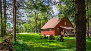 Une cabane en bois rouge, dotée d’un porche, entourée d’une cour verdoyante et d’arbres.