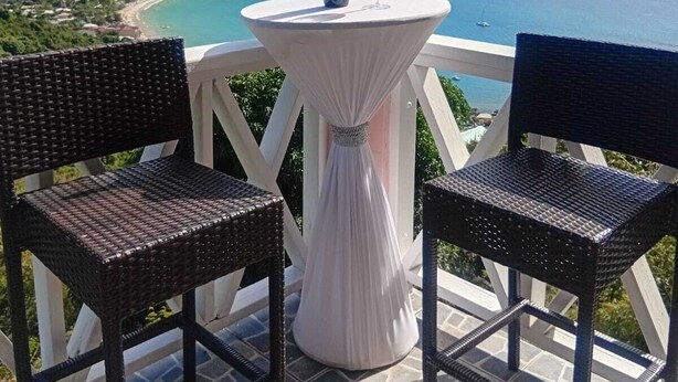 Un balcon agrémenté de fauteuils en osier, une table avec une bouteille de vin et un verre, offrant une vue sur un paysage côtier avec des montagnes et une plage.