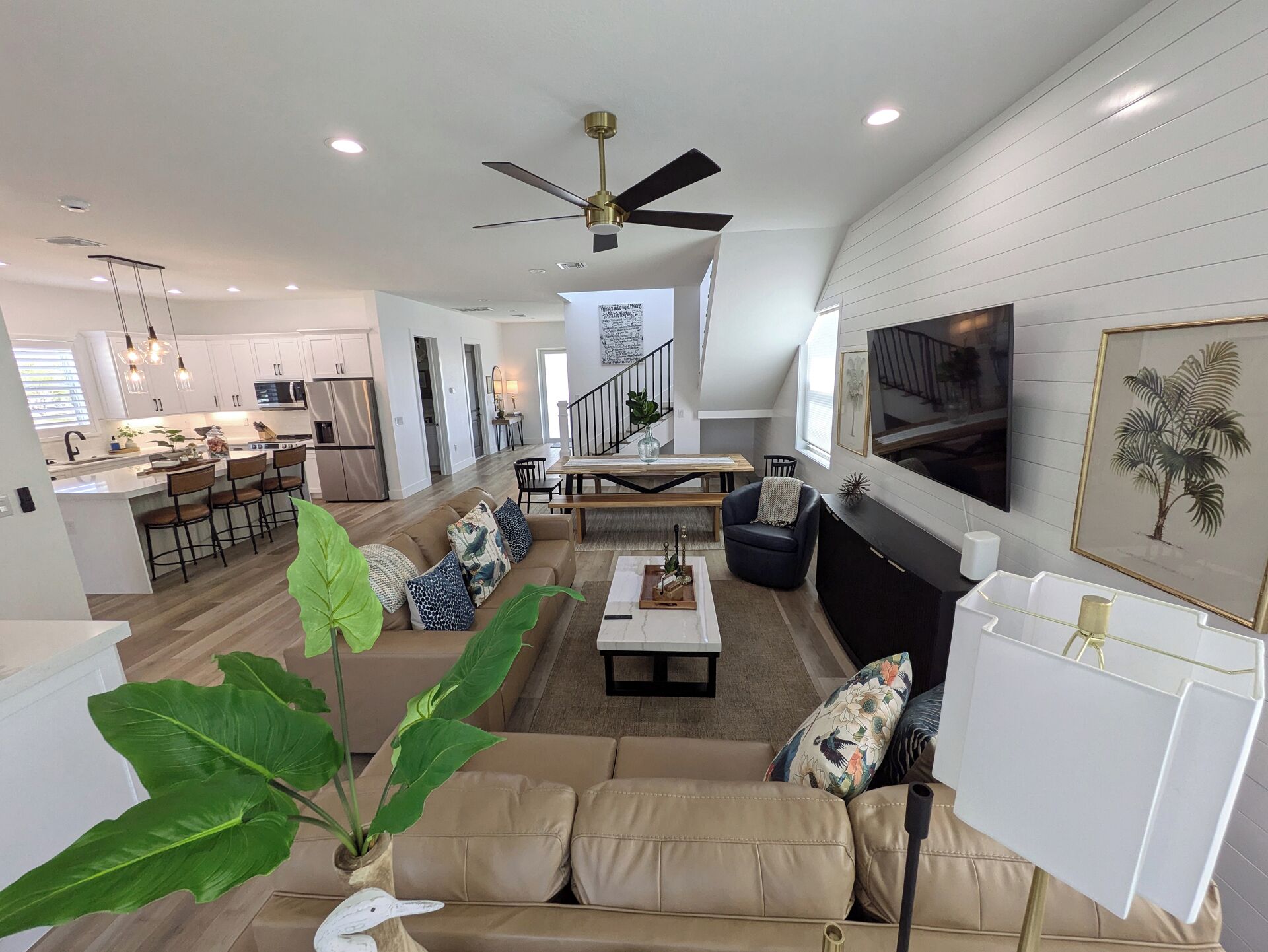 A modern living room with a sofa, coffee table, and a TV mounted on the wall. There is a kitchen area in the background with a dining table and chairs.