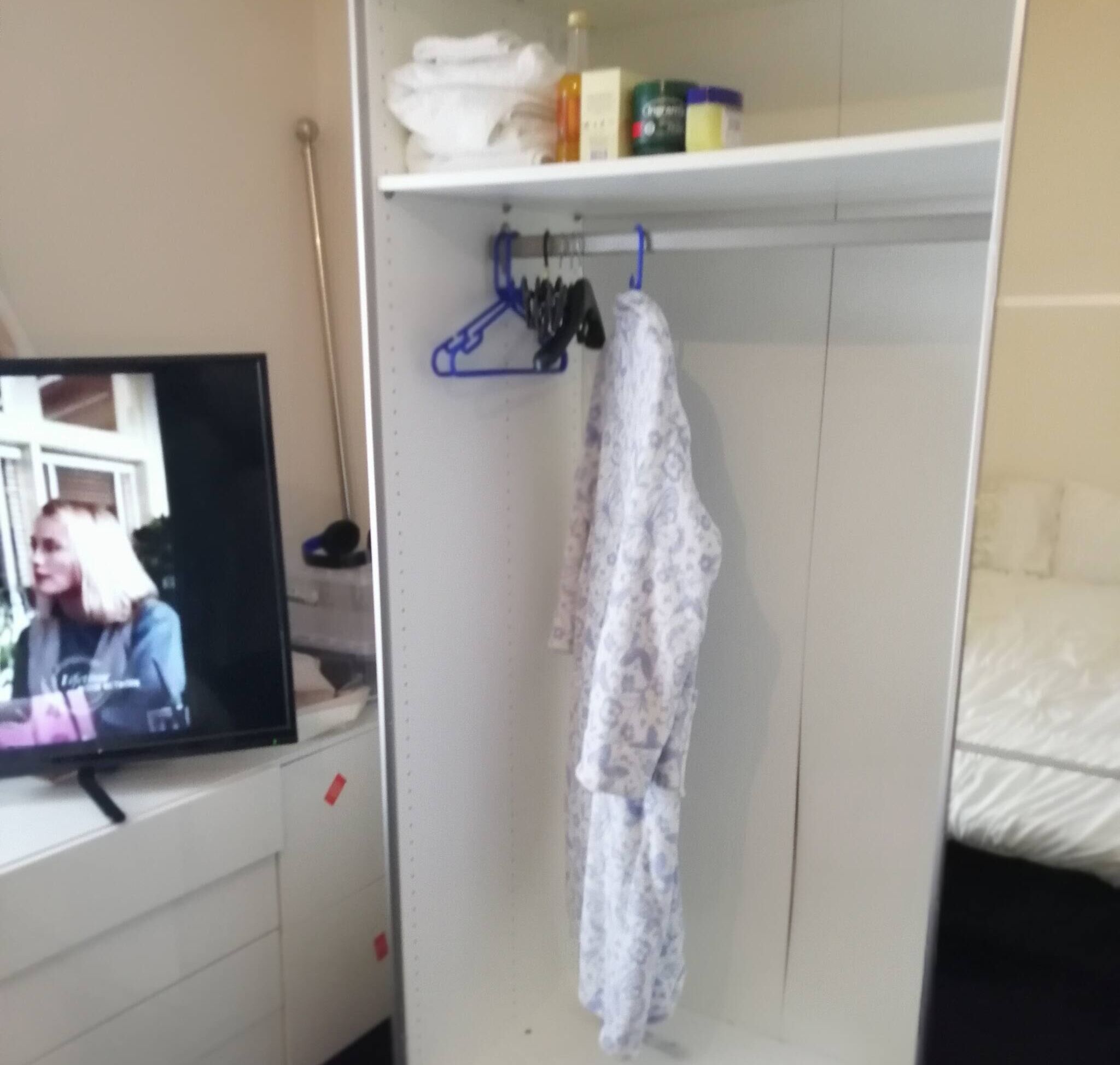 Una stanza con televisione accesa su cui si vede una persona, un armadio con abiti appesi e uno scaffale con articoli da toilette.