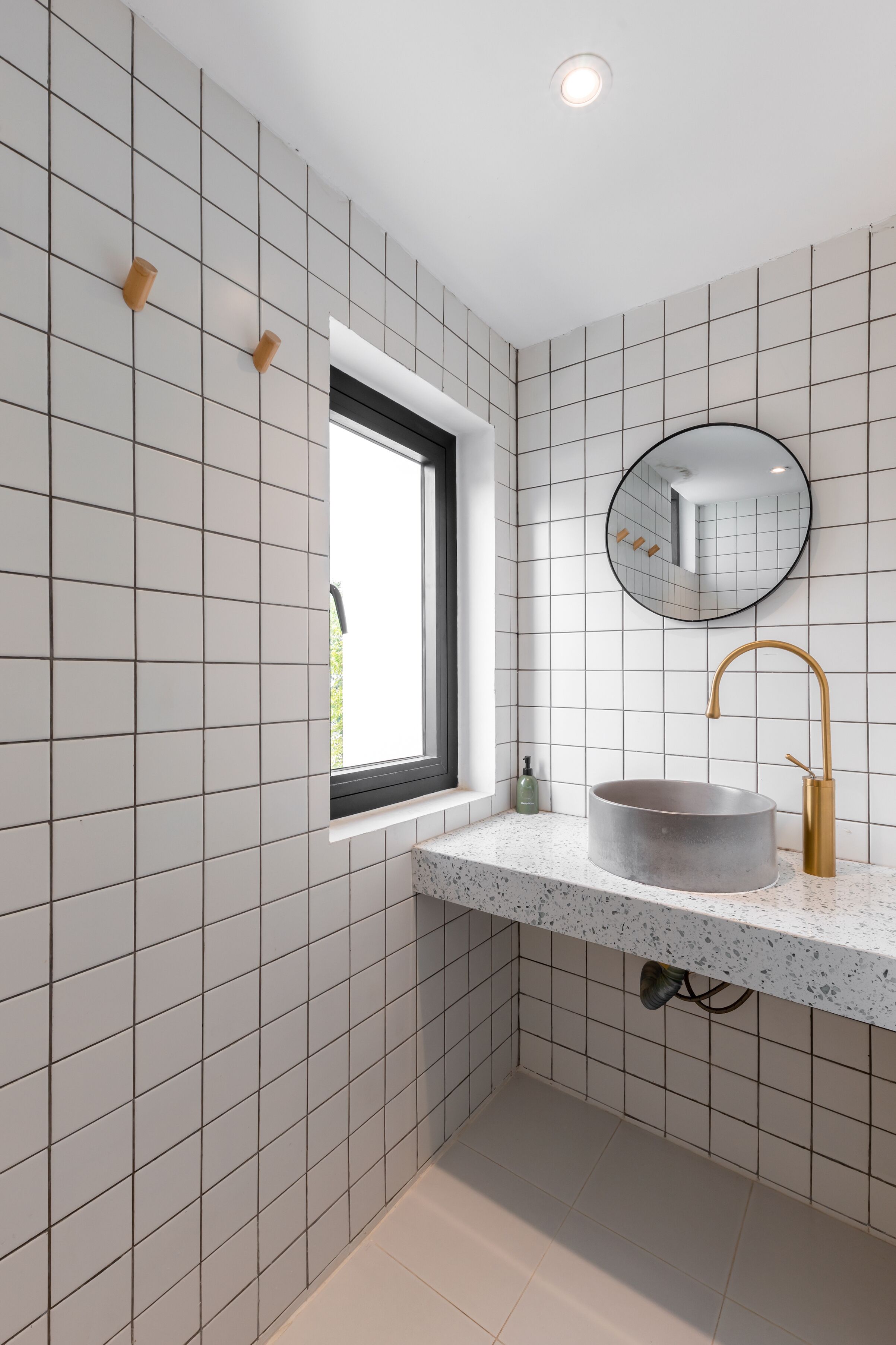 A bathroom with white tiled walls, a round mirror, a round sink with a gold faucet, and a window with a black frame.