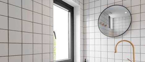A bathroom with white tiled walls, a round mirror, a round sink with a gold faucet, and a window with a black frame.