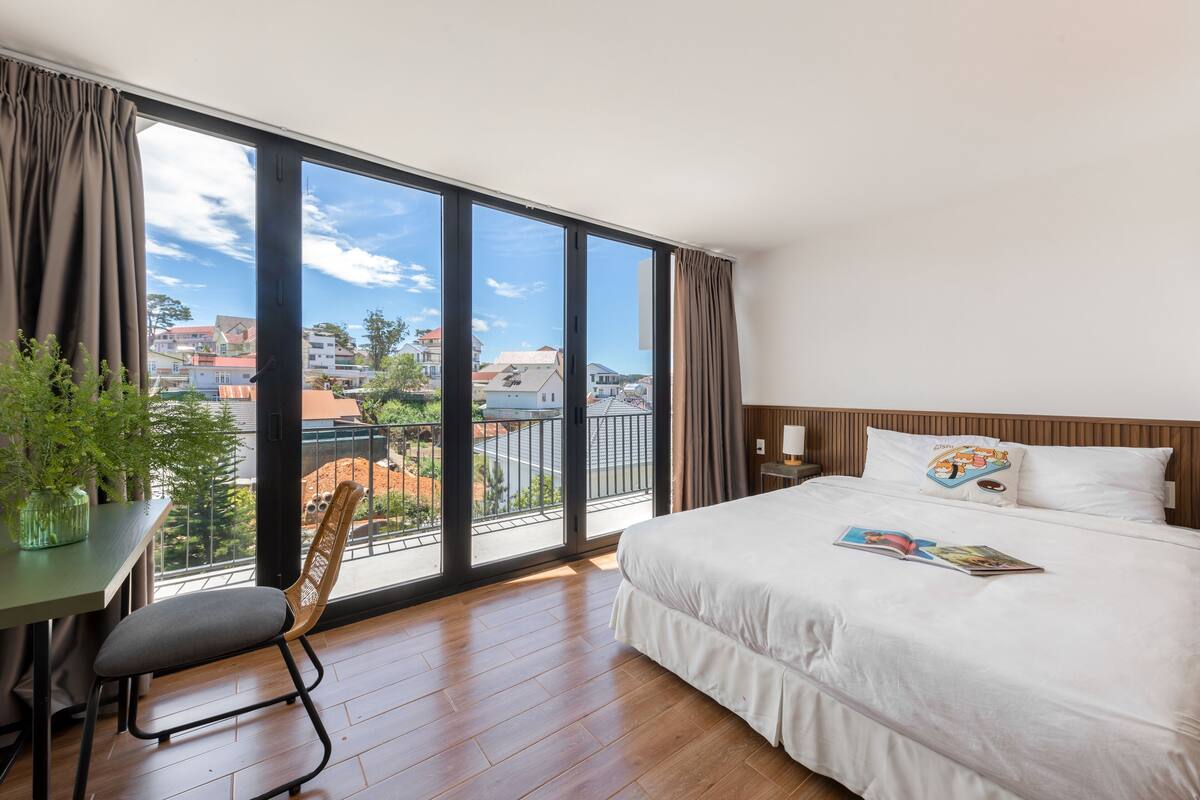 A hotel room with a large window, a bed, a desk with a chair, and a potted plant.