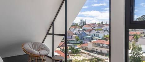 Una habitación con techo inclinado, un ventanal con vistas a edificios, una silla de mimbre, un taburete pequeño y un tapete estampado.