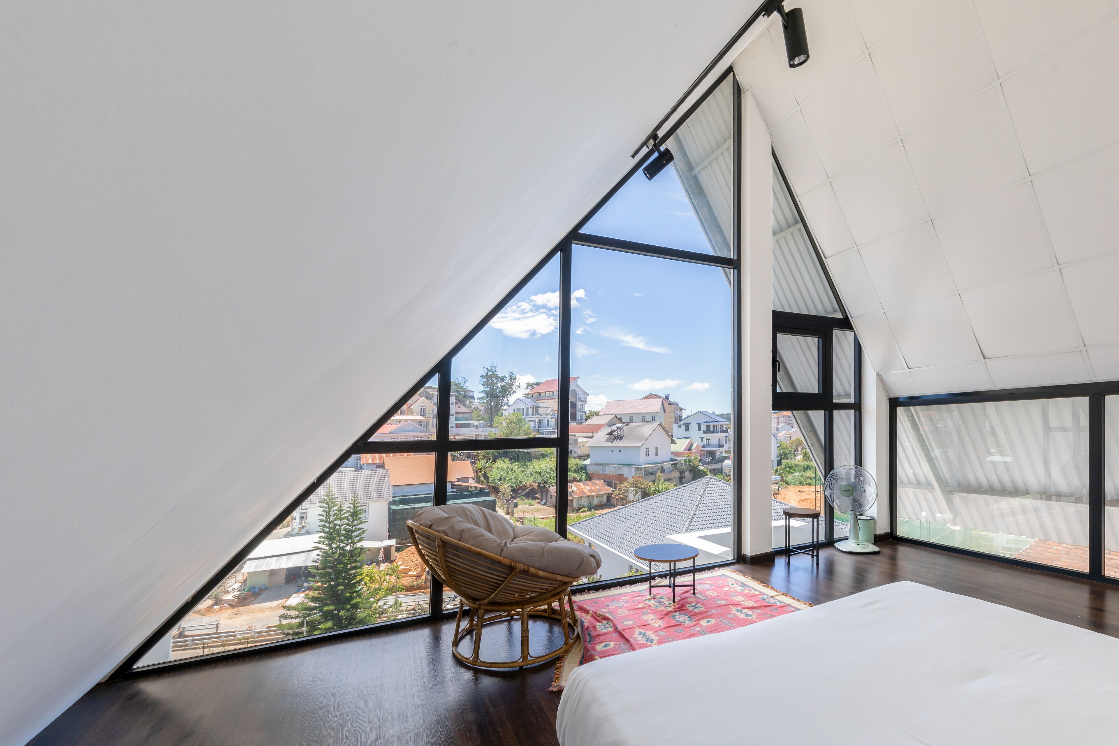 A bedroom with a large window, a bed, a wicker chair, and a small table.